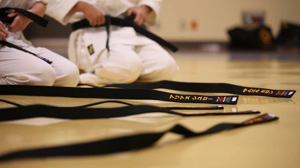 Karateka berlutut di lantai dojo dengan deretan sabuk hitam bersulam nama di depannya