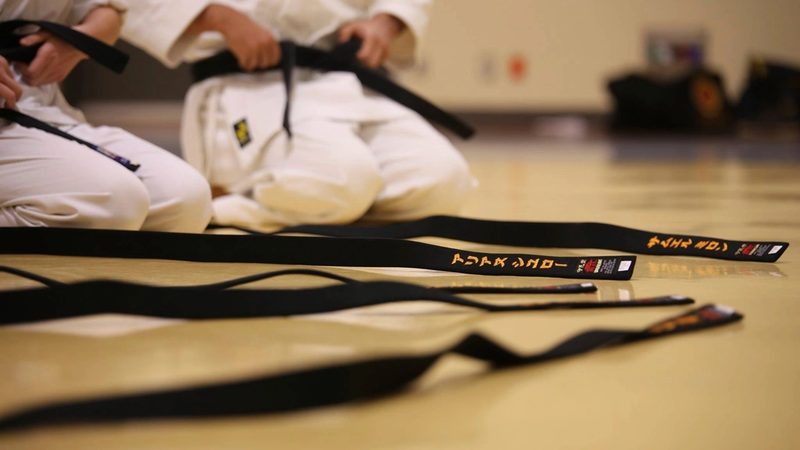 Karateka berlutut di lantai dojo dengan deretan sabuk hitam bersulam nama di depannya